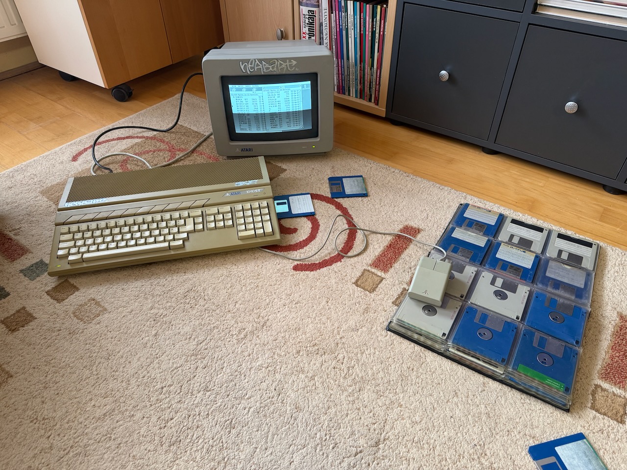 An Atari STE on the carpet in my living room, connected to an Atari SM124 monochrome monitor. To the right is a folder of floppy disks that also serves as a mouse pad.